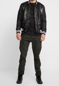Veste bomber en cuir noir avec poignets à rayures blanches, associée à une chemise texturée gris foncé et un pantalon cargo vert olive. Baskets noires.