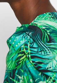 Top con stampa tropicale caratterizzato da motivi verdi e blu, con dettagli di spalle arricciati e una texture di tessuto liscia.