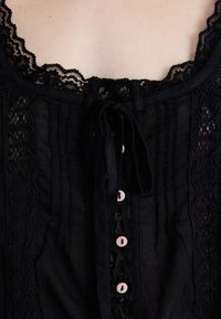 Ingrandimento di un indumento nero con pizzo e bottoni rosa, con un fiocco legato al colletto, che mostra dettagliate trame del tessuto.