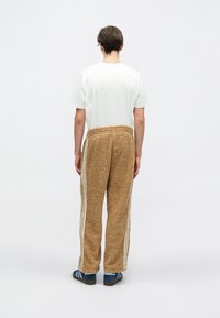 Pantalones marrones texturizados con rayas laterales blancas, combinados con una camiseta blanca sencilla. Usados con zapatillas de deporte con rayas azules. Vista posterior.