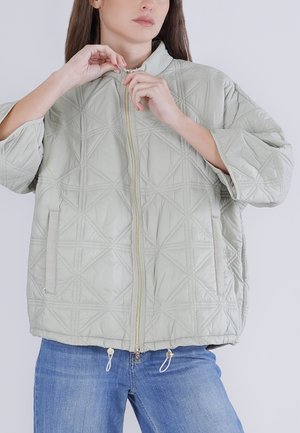 Kvinde iført lysegrå quiltet jakke med geometrisk syning, delvist lynlåst, og blå jeans, foran en ensfarvet baggrund.