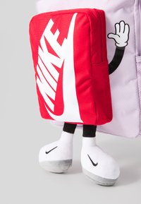 Rdeča tekstilna torbica z belim logotipom Nike, pritrjena na pastelno vijoličastem nahrbtniku. Ima črne roke in puhaste bele-sive čevlje spodaj.