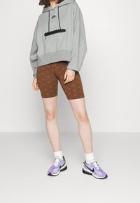 Sudadera corta gris con cordones negros, pantalones cortos de bicicleta marrones con patrón de logotipo repetido, calcetines blancos de caña larga y zapatillas moradas y negras.