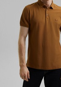 Polo shirt marron en tissu texturé, avec une patte de boutonnage à deux boutons et un petit logo sur la poitrine gauche. Manches courtes, design ajusté.