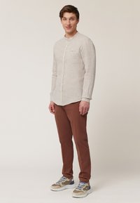 Lichtbeige linnen overhemd met een staande kraag en knoopsluiting, gecombineerd met roestkleurige broek en beige sneakers met blauwe accenten.