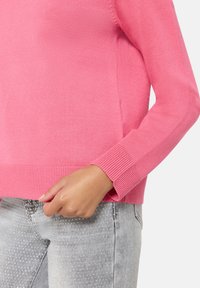 Personne portant un pull à manches longues rose vif et un jean gris clair avec des motifs à pois blancs, la main tenant légèrement l'ourlet du pull.