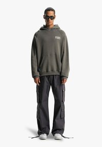 Felpa oversize verde scuro con tasca frontale e logo "PEQUS". Abbinata a pantaloni cargo neri con più tasche. Sneakers bianche e occhiali da sole.