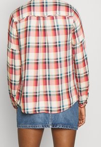 Geruite shirt van zachte stof met rode, blauwe en crèmekleurige tinten. Het ontwerp heeft lange mouwen en een afgeronde zoom. Jeansrok zichtbaar eronder.
