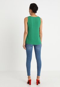 Kvinna som står med ryggen synlig, klädd i en ärmlös grön topp, blå skinny jeans och röda högklackade skor mot en vit bakgrund.