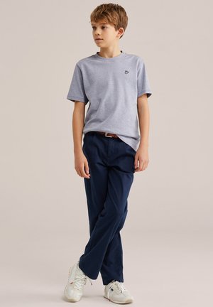 Niño de pie con las piernas cruzadas, vestido con una camisa de manga corta color gris claro, pantalones azul marino con cinturón y zapatillas blancas, mirando hacia un lado.