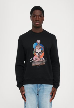 Young man wearing a black sweatshirt with a colorful snake, skull design, and "Coney Island Cobras" text, paired with light blue jeans.