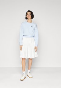 Light blue sweatshirt with embroidered text, paired with a white pleated skirt and white sneakers with gum soles. Clean, casual look.