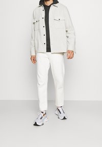 Giacca button-up grigia chiara sopra un felpa scura, abbinata a pantaloni bianchi e sneakers grigie. Caratteristiche includono tasche e texture contrastanti.