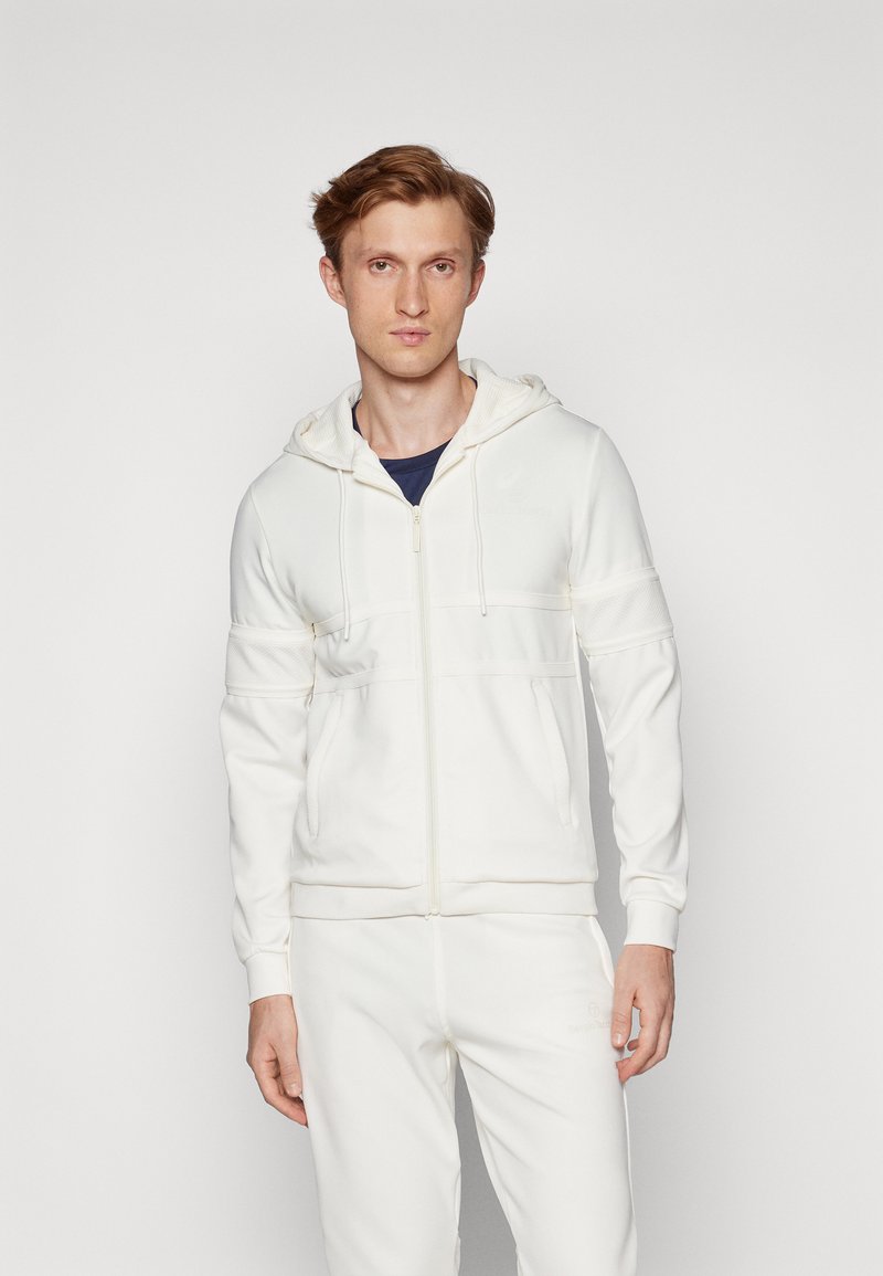 Hvit zip-up hettegenser med teksturerte paneler, snøring på hetten og to frontlommer, kombinert med matchende joggebukser i et mykt stoff.