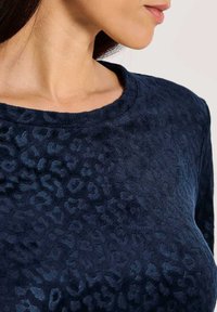 Vrouw in een marineblauw shirt met een subtiele textuur van luipaardprint, waarbij haar nek, schouder en een deel van haar gezicht in profiel zichtbaar zijn.