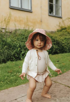 Bambino con cappello rosa, cardigan bianco e abbigliamento beige, in piedi a piedi nudi su un sentiero di pietra vicino a cespugli verdi.