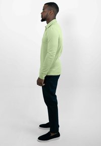 Heldergroene poloshirt met lange mouwen en een kraag, gecombineerd met donkere broek en zwarte instappers, gezien vanaf de zijkant.