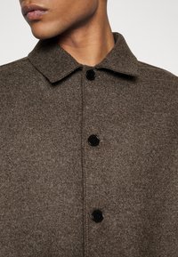 Manteau en laine mélangée marron avec une finition texturée, présentant un col, des boutons noirs et une coupe droite, mettant en avant des lignes épurées et un design minimal.