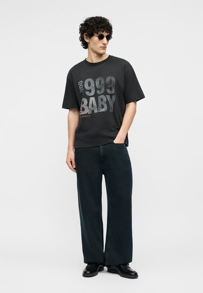 Άνδρας με σγουρά μαλλιά και γυαλιά ηλίου, φορώντας μαύρο υπερμεγέθες T-shirt με την επιγραφή "999 BABY", σκούρα παντελόνια φαρδιάς γραμμής και μαύρα παπούτσια.