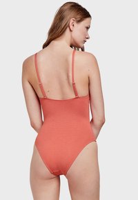 Maillot de bain une pièce en corail, en tissu texturé avec de fines bretelles réglables, design dos nu et ouvertures de jambes échancrées.