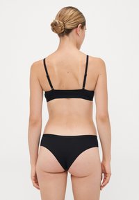 Woman with braided bun hairstyle wearing a black bra and matching bikini-style underwear, photographed from behind on a plain background.