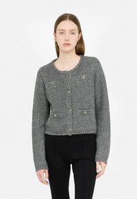 Cardigan grigio a maglia con scollo rotondo, frontale con bottoni e due tasche applicate con decorazioni. Indossato sopra pantaloni neri.