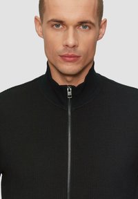 Schwarzer Reißverschluss-Pullover mit hohem Kragen. Der strukturierte Stoff weist gerippte Abschnitte und einen glatten Reißverschluss auf. Enganliegendes Design, keine Muster oder Akzente.