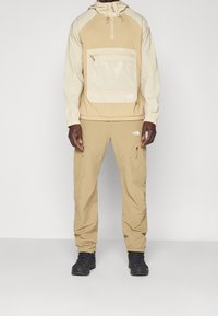 Chaqueta beige ligera con un gran bolsillo de malla, capucha ajustable y puños elásticos, combinada con pantalones a juego y botas de senderismo negras.