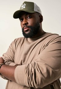 Khaki cap with gray mesh back, featuring white logo. Light brown long-sleeve shirt with relaxed fit, arms crossed, showcasing bracelet.