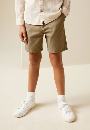 Bambino che indossa pantaloncini beige, scarpe da ginnastica bianche, calzini bianchi e una maglietta beige chiaro a maniche lunghe, in piedi contro uno sfondo neutro.