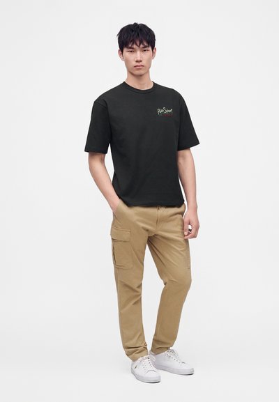 Νέος άνδρας που φοράει μαύρο t-shirt Polo Sport, καφέ παντελόνι τύπου cargo και λευκά αθλητικά, στέκεται με το ένα χέρι στην τσέπη σε λευκό φόντο.