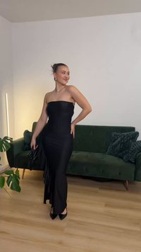 Črna večerna obleka brez naramnic, oprijet kroj, rahlo razširjen rob, gladek material, dopolnjena z majhno torbico, obuta v čevlje s koničastim peto.