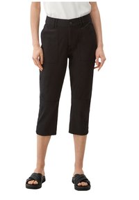 Donna che indossa pantaloni neri cropped con grandi tasche frontali, una canotta bianca e sandali neri a scivolo su uno sfondo bianco.