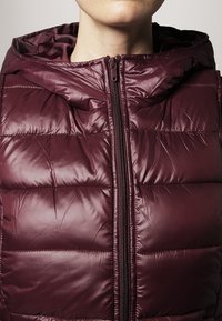 Bordeauxrote Steppweste mit glänzender Textur, Reißverschluss und Kapuze. Verfügt über horizontale Steppnähte zur Isolierung.