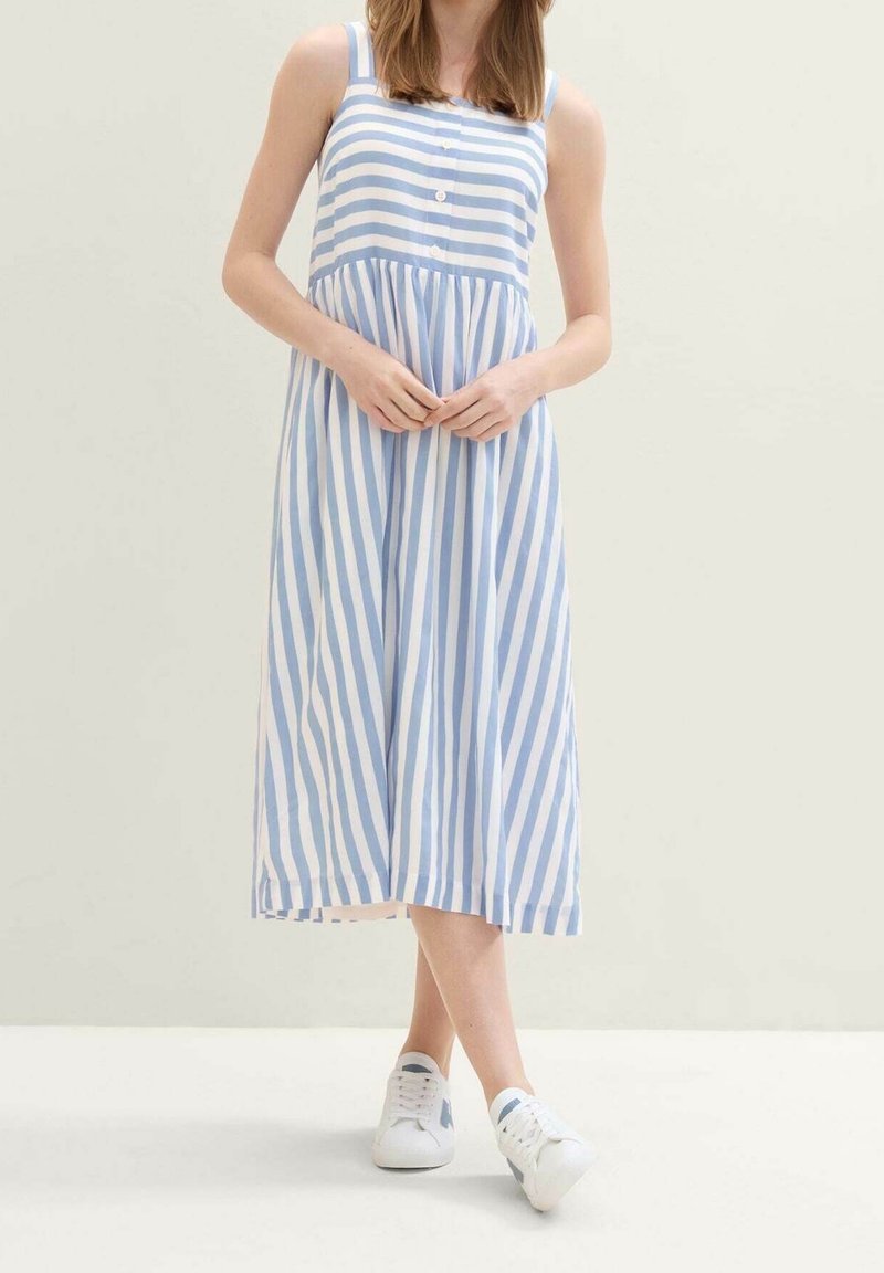 Femme portant une robe midi sans manches à rayures verticales bleues et blanches et des baskets blanches, debout les mains jointes.