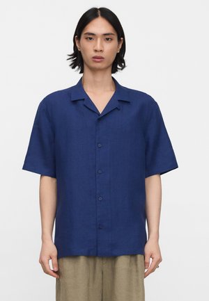 Jong persoon met zwart haar tot op de schouders, met een loszittend, donkerblauw overhemd met korte mouwen en een beige broek, staand tegen een witte achtergrond.