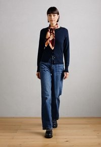 Navy cardigan over blue wide-leg jeans, paired with a patterned silk scarf in red, orange, and cream, and black lace-up shoes.
