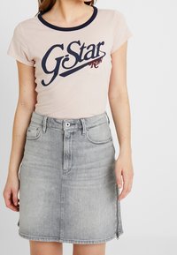Jasnoróżowa koszulka G-Star z granatowym dekoltem typu crew i dużym nadrukiem logo. Szara spódnica dżinsowa z przednim zamkiem błyskawicznym i pięcioma kieszeniami, długość powyżej kolan.