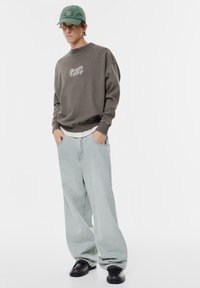 Szürke pulóver "Death Valley" felirattal, világoskék, bővülő farmernadrággal és fekete cipővel. A zöld baseball sapka egészíti ki az összeállítást.