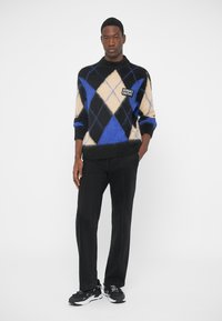 Svart och blå argylemönstrad stickad tröja med gräddvita diamantmönster, med en logotyp. Matchas med svarta byxor och svarta sneakers.