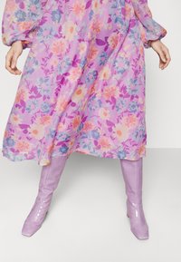 Vestido floral com padrões roxos, cor-de-rosa e azuis; tecido leve, mangas longas. Combinado com botins brilhantes até ao joelho em lavanda.