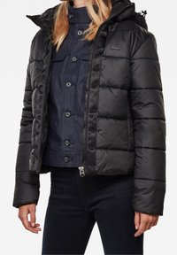 Černá péřová bunda se zipem vpředu, kapucí a žebrovanými manžetami. Má lesklý povrch, prošívaný vzor a je nošena přes tmavou džínovou košili.