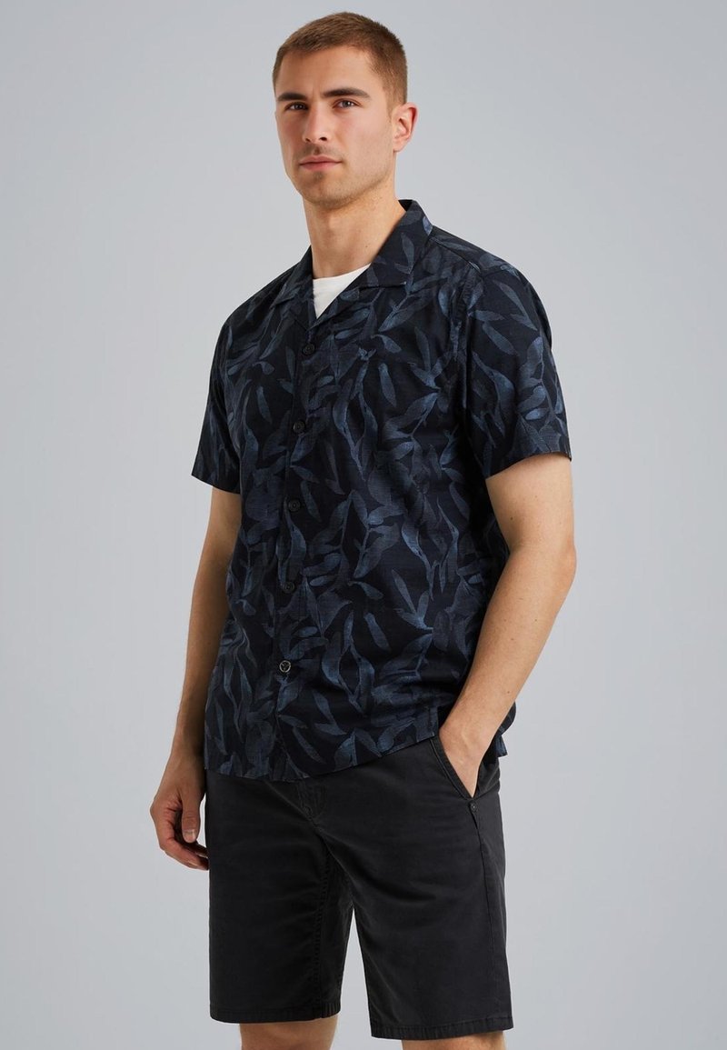 Camisa negra de manga corta con patrón de hojas azul oscuro, diseño de botones, cuello y tejido ligero. Combinada con pantalones cortos negros.