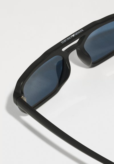 Melnas aviator stila saulesbrilles ar matētu apdari, zilu tonētu lēcu un modernu ģeometrisku dizainu. Liktas sudraba emblēmas uz rāmja.
