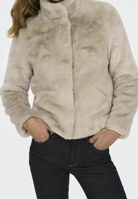 Ljus beige faux fur-jacka med hög krage och långa ärmar, med en mjuk textur och minimalistisk design, i kombination med mörka jeans.