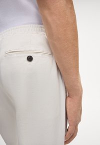 Witte, lichte broek met een elastische tailleband. Voorzien van een achterzak met een donkere knoopdetail. Gladde textuur, slim-fit snit.