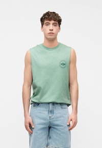 Νέος άνδρας με πράσινο αμάνικο τοπ με μικρό λογότυπο surf και ανοιχτόχρωμα τζιν σορτς, στέκεται μπροστά σε λευκό φόντο.