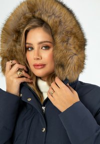 Granatowa kurtka puchowa z dużym, teksturowanym kapturem z sztucznego futra. Posiada zamek błyskawiczny i złote akcenty. Pod spodem miękki kremowy golf.