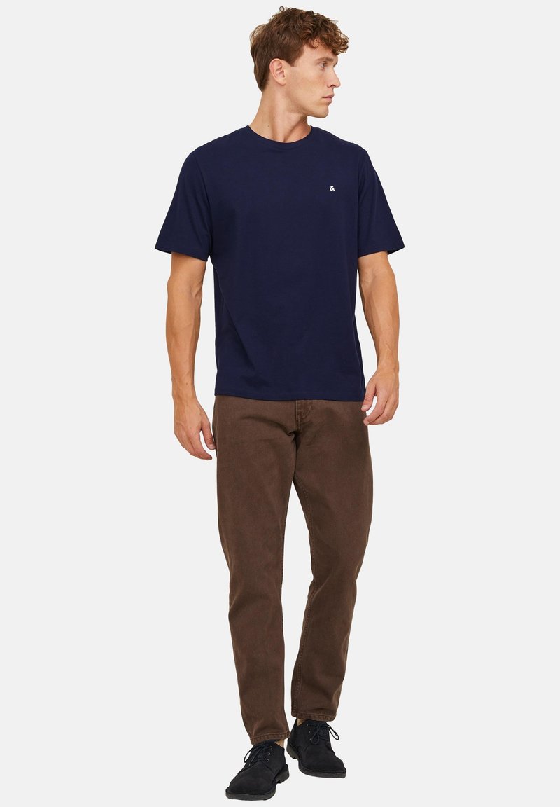 Homme portant un t-shirt bleu marine à manches courtes et un pantalon marron, debout et regardant vers sa gauche sur un fond blanc.