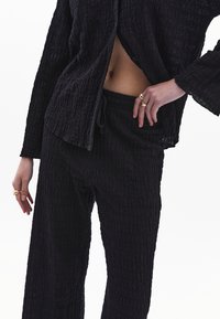 Černá souprava s texturou zahrnující volný, oversized top s knoflíky a kalhoty na šňůrku, obojí s mačkanou úpravou látky.